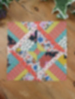 A colorful patchwork hidden wells quilt block on a wooden surface, featuring geometric patterns and floral prints in orange, pink, yellow, and blue.