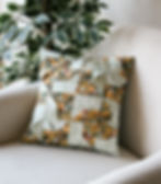 Floral patchwork pillow on beige armchair, green leafy plant in background. Vibrant patterns with white, yellow, and orange flowers. Cozy setting.