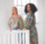 Two women in floral outfits smile by a white railing in a bright room with large windows and a plant. One wears green, the other orange.