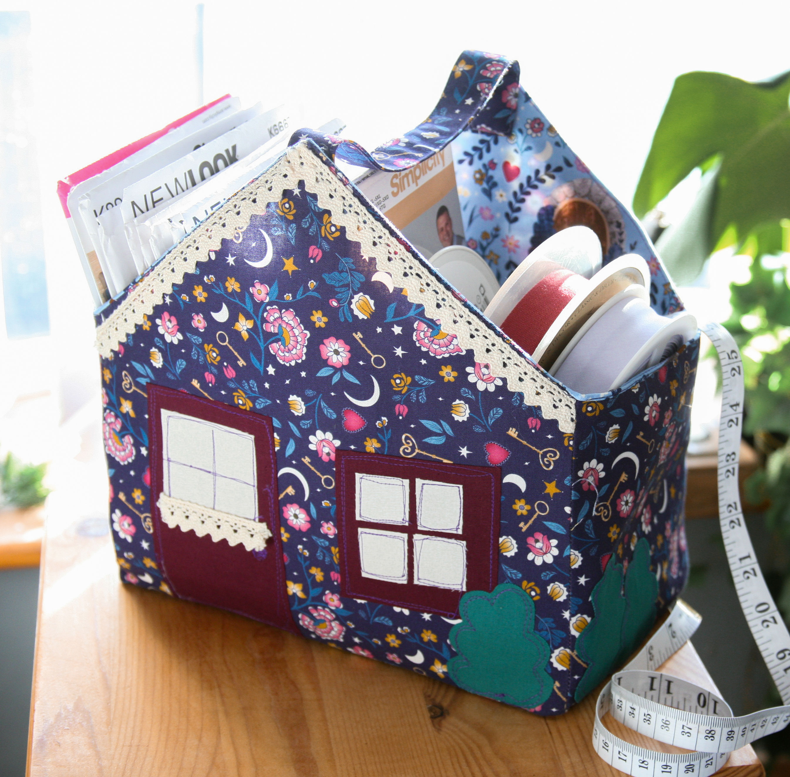 the cottage fabric basket in purple whimsical fabrics, sewing patterns are stored inside and it is on a wooden table top next to plants