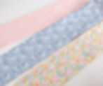 Three strips of fabric with floral patterns; pink with dots, blue with flowers, and multicolored with fruit and bees on a white surface.