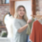 Smiling woman in a sewing room with a mannequin, holding a green garment on a rack. Brick wall and flowers in the background.