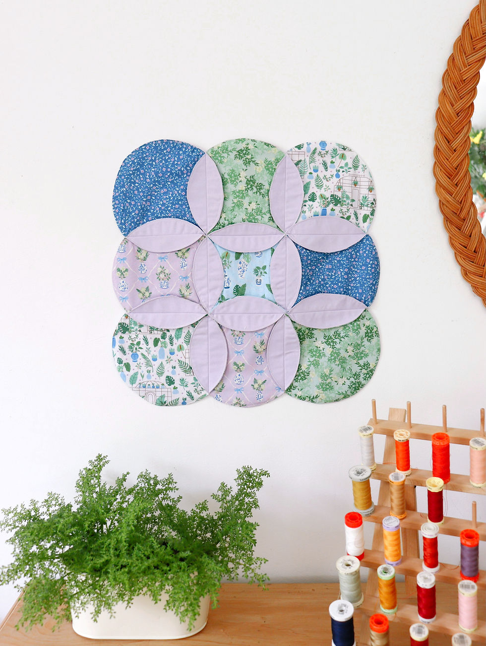 Patchwork circles with blue and green floral patterns on a white wall. A fern in a pot and colorful thread spools on a wooden shelf below.