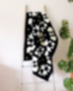 Black-and-white geometric quilt draped over a wooden ladder against a white wall. A green leafy plant is visible on the right side.