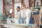 Two women in a cozy sewing room, one adjusts the other's sleeve. A sewing machine and fabrics sit on a wooden table. Brick wall backdrop.