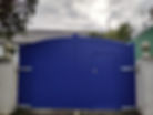 Solid blue timber driveway gate with curved top and pedestrian access door, installed between concrete piers