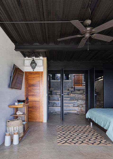 Modern bedroom at Casa Zenta featuring a cozy king-sized bed, minimalist decor, ceiling fan, and a wall-mounted TV. The room offers direct access to an outdoor space through a wooden door, leading to a tranquil garden view, ideal for a peaceful retreat in the heart of nature.