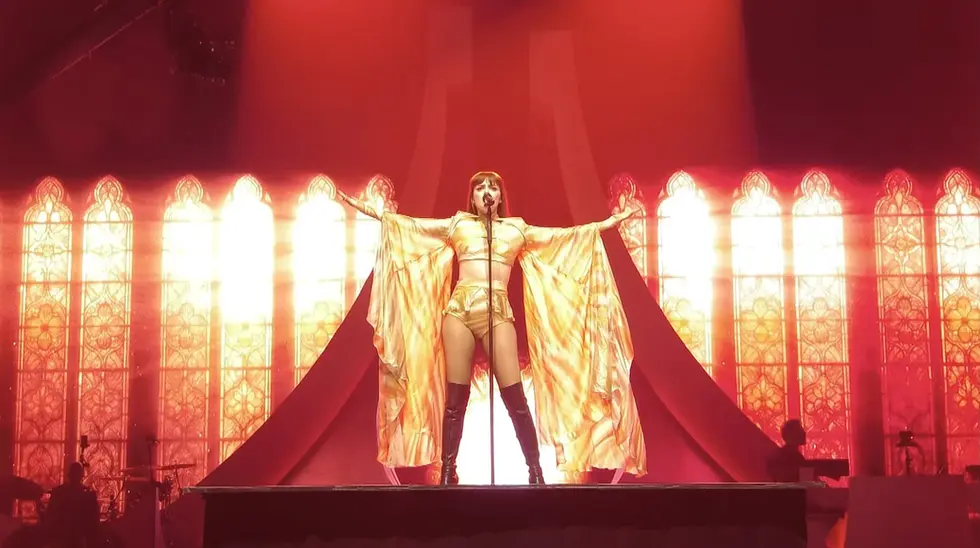Annalisa a braccia aperte sul palco con un costume dorato scintillante e mantello, davanti a uno schermo con grafiche di vetrate luminose.