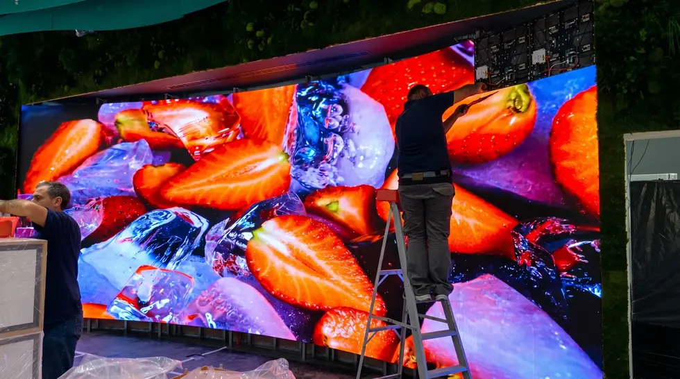 Fase di installazione tecnica di un display LED curvo Lamina SR all'interno di uno store, con lo schermo acceso che visualizza un dettaglio macro ad alto contrasto di fragole e cubetti di ghiaccio.