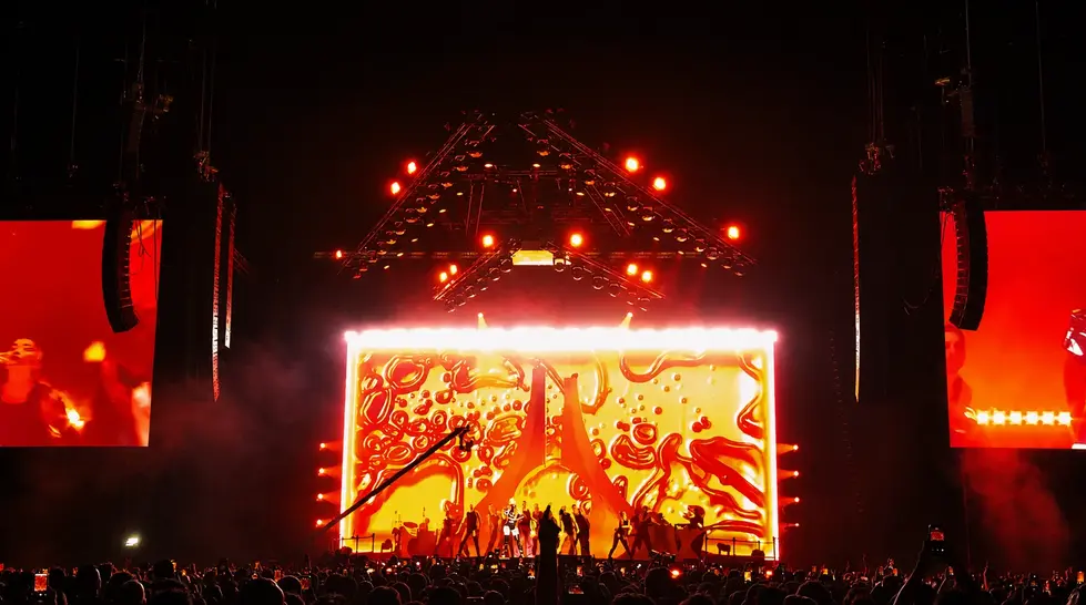 Ampia visuale di un concerto pop con grandi schermi arancioni, strutture di luci a soffitto e silhouette del pubblico in primo piano.