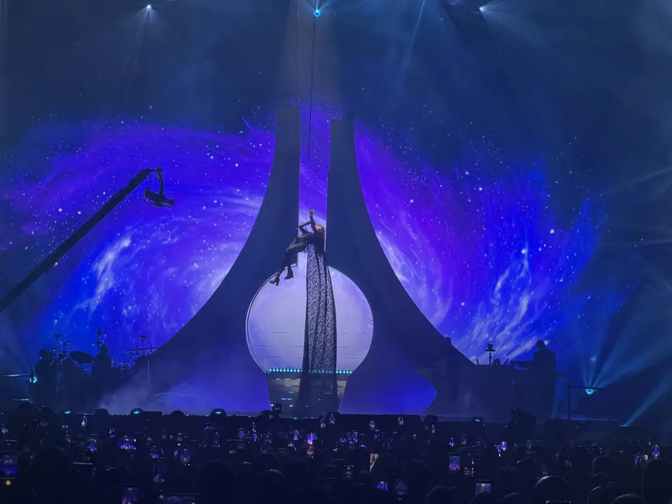Cantante sospesa in aria al centro di una struttura architettonica illuminata su uno sfondo di una nebulosa spaziale viola e blu durante un concerto.
