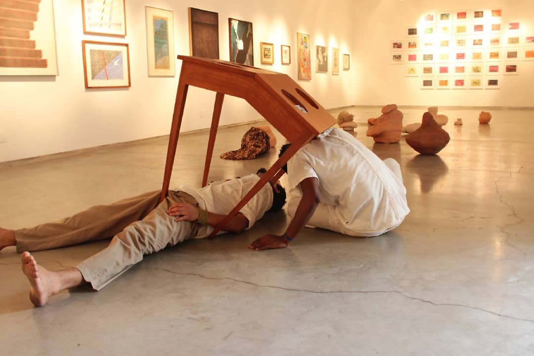 An action in the exhibition space, with two people beside a tilted table and artworks on the walls and floor