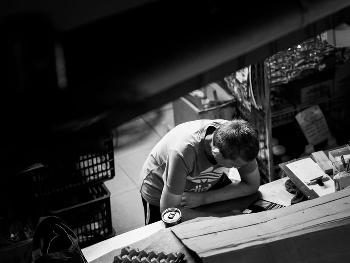 Man leaning forward over a counter with papers, cartons, and a drink can in a dim interior workspace, photographed by Gary PWK for the Made In His Image project.