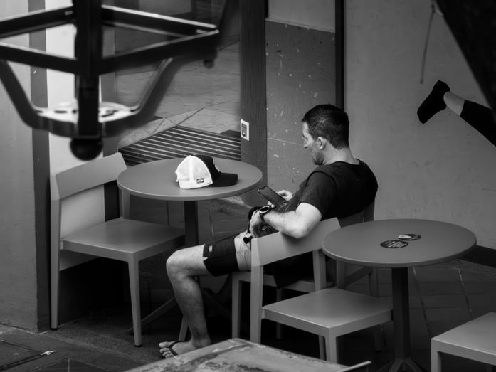 A man sits alone at a small table in a shaded cafe area, looking down at his phone, with empty chairs and tables around him, photographed by Gary PWK for the Made In His Image project.