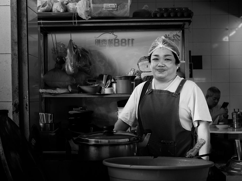 A woman wearing an apron stands behind a kitchen counter inside a family dining space, with prepared food and containers nearby before a gathering begins, photographed by Gary PWK for the Made In His Image project.
