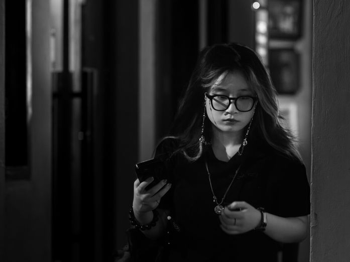 Woman walking through a dim corridor while holding a phone, light falling across the passageway in an evening street scene, photographed by Gary PWK for the Made In His Image project.