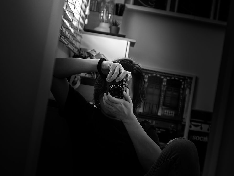 GaryPWK sits indoors holding a camera up to a mirror, partially obscuring their face in a quiet room, photographed by Gary PWK for the Made In His Image project.