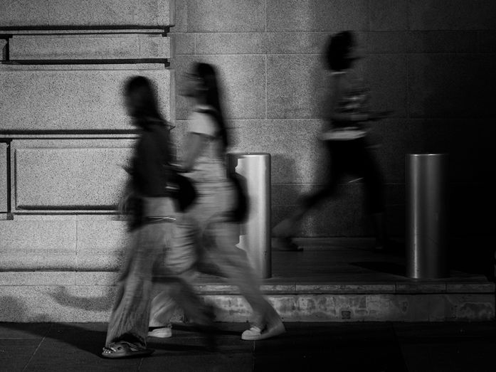 Blurred figures walking past a stone wall, suggesting movement, time, and the quiet weight of what remains carried forward.