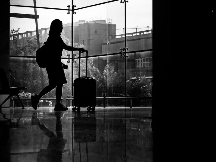 Silhouette of a traveler standing in an airport terminal beside a suitcase, looking through large windows toward an aircraft outside, reflective travel scene.