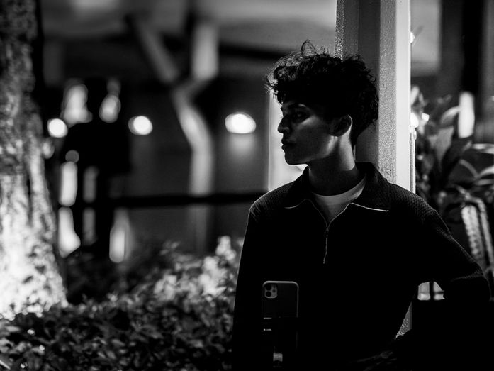 A young man stands alone at night beside a pillar in an urban setting, facing sideways with a still posture as lights blur softly in the background, photographed by Gary PWK for the Made In His Image project.
