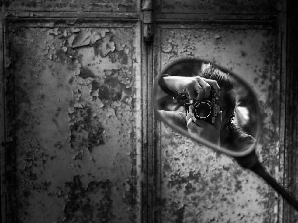 Gary holding a camera is seen through a small oval mirror mounted on a weathered, peeling wall surface, photographed by Gary PWK for the Made In His Image project.