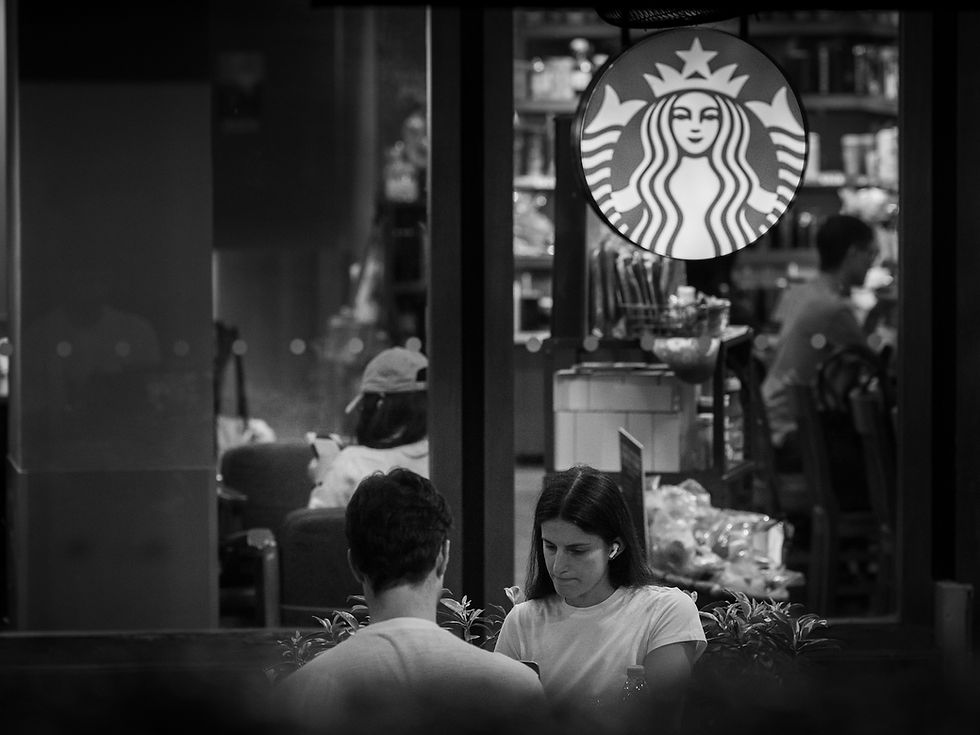 Two people seated outside a café at night, seen through a black and white street scene along Orchard Road, photographed by Gary PWK for the Made In His Image project.