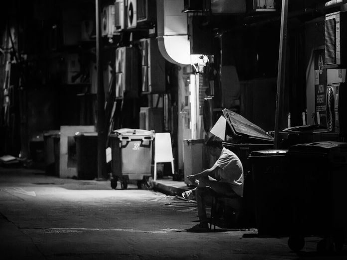 Black and white urban street scene showing a solitary man resting in a narrow alleyway at night under harsh lighting, representing unnoticed strength and everyday human resilience.