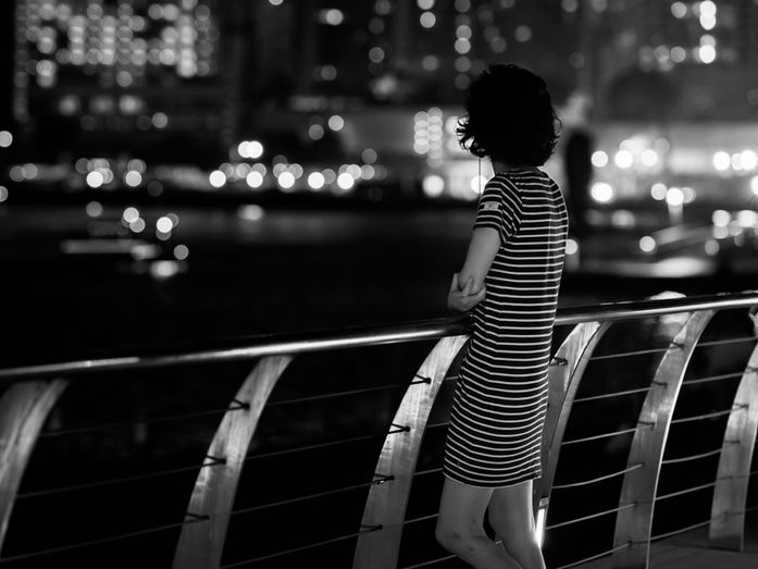A woman stands alone at a railing at night, looking out over softly glowing city lights, evoking quiet presence and a season of uncertainty.
