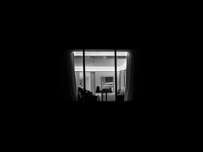 A brightly lit apartment interior is seen through a large window at night, with curtains drawn open and furniture visible inside against the surrounding darkness, photographed by Gary PWK for the Made In His Image project.