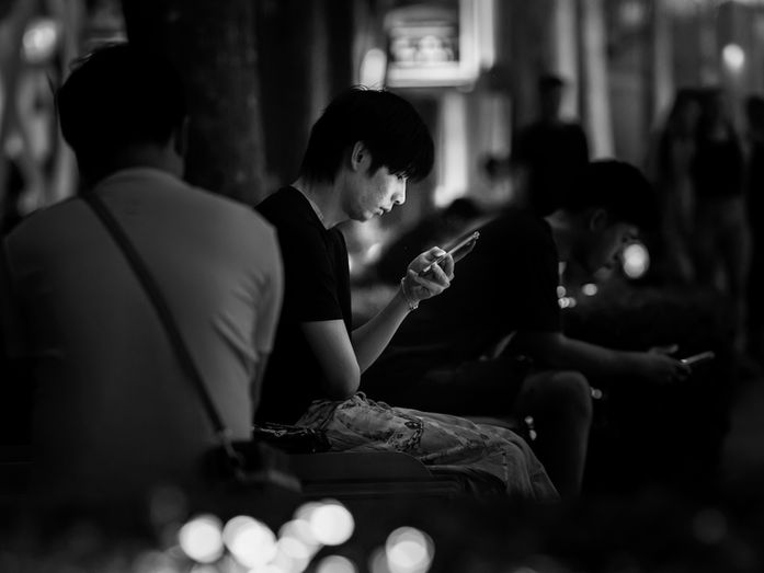 A young man sitting on a bench in a dimly lit public space looking at his phone while others sit nearby, photographed in Singapore for the Made In His Image project, photographed by Gary PWK for the Made In His Image project.