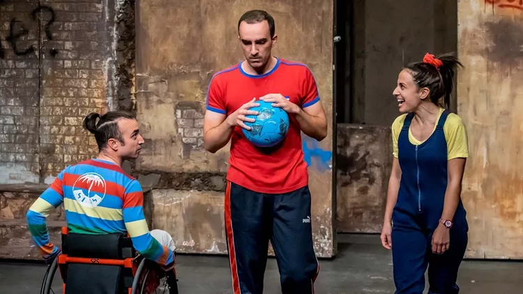 On the right, Zili with an intense expression of joy. On the left, Aris in a wheelchair. Both are looking at Fanis in the middle, who is holding a blue ball while looking thoughtful. They are all are wearing brightly colored clothing.