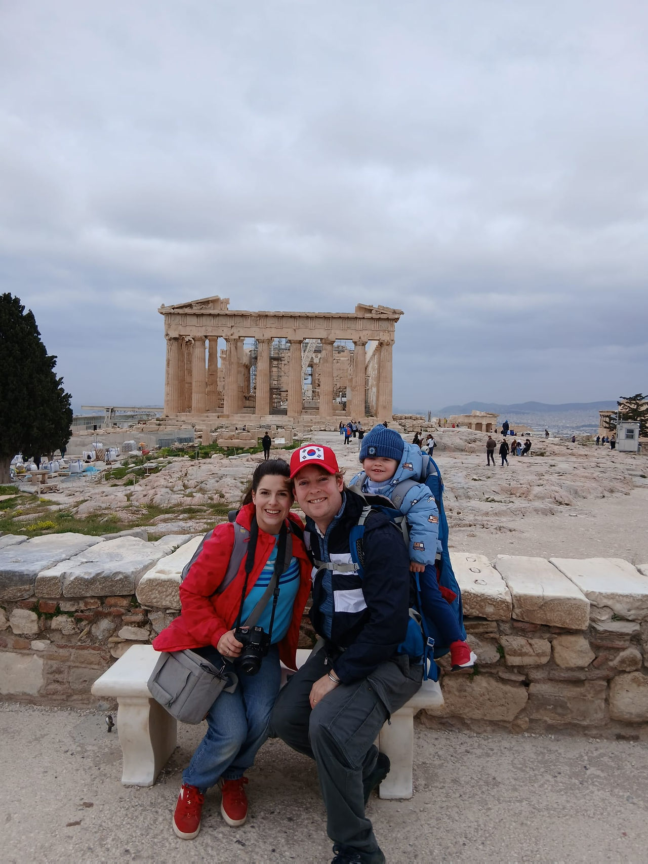 A smiling family of three sit on a bench with the Parthenon behind them. The father is carrying the toddler in a backpack carrier.