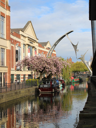 A beautiful river view in Lincoln, blue sky and spring flowers. Empowerment, a steel statue of two figures stretching out to each other across the water, a pink blossom tree is in the foreground.