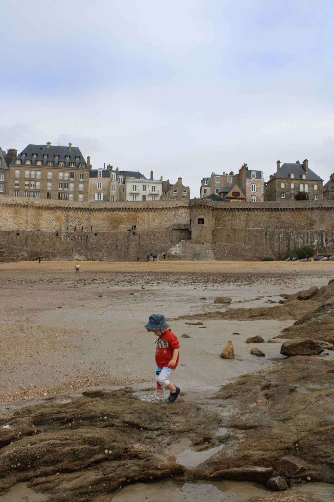 A very young boy in a orange jumper, grey shorts, and a bucket hat walks across a rocky, wet beach at low tide. In the background, the massive stone ramparts of Saint-Malo, France, rise up from the sand, topped by historic multi-story buildings under a cloudy sky.