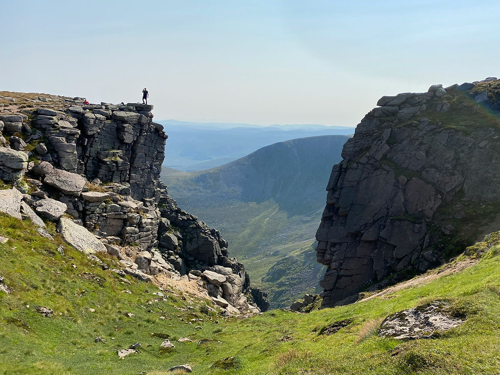 Spectacular views at Cairngorms National Park. A tiny hike looks from a rocky crag down into a green valley, with Scottish mountains in the distance.