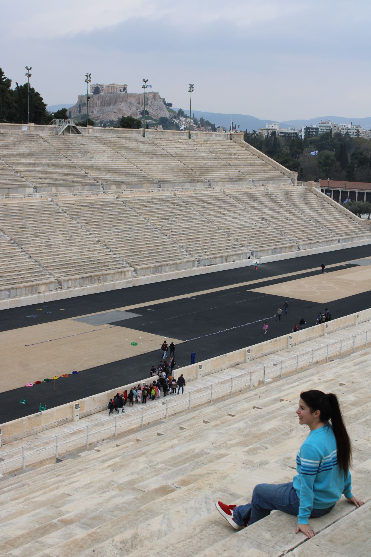 A high-angle view looking down into the massive, empty Panathenaic Stadium in Athens. Built entirely of white marble, the steep, multi-tiered seating rows cascade towards the modern black running track. A woman in a blue jumper sits on the stone seats in the foreground, looking over the arena. Rising in the distance, centered behind the stadium, is the ancient Acropolis hill with the columns of the Parthenon clearly visible against the cloudy sky.