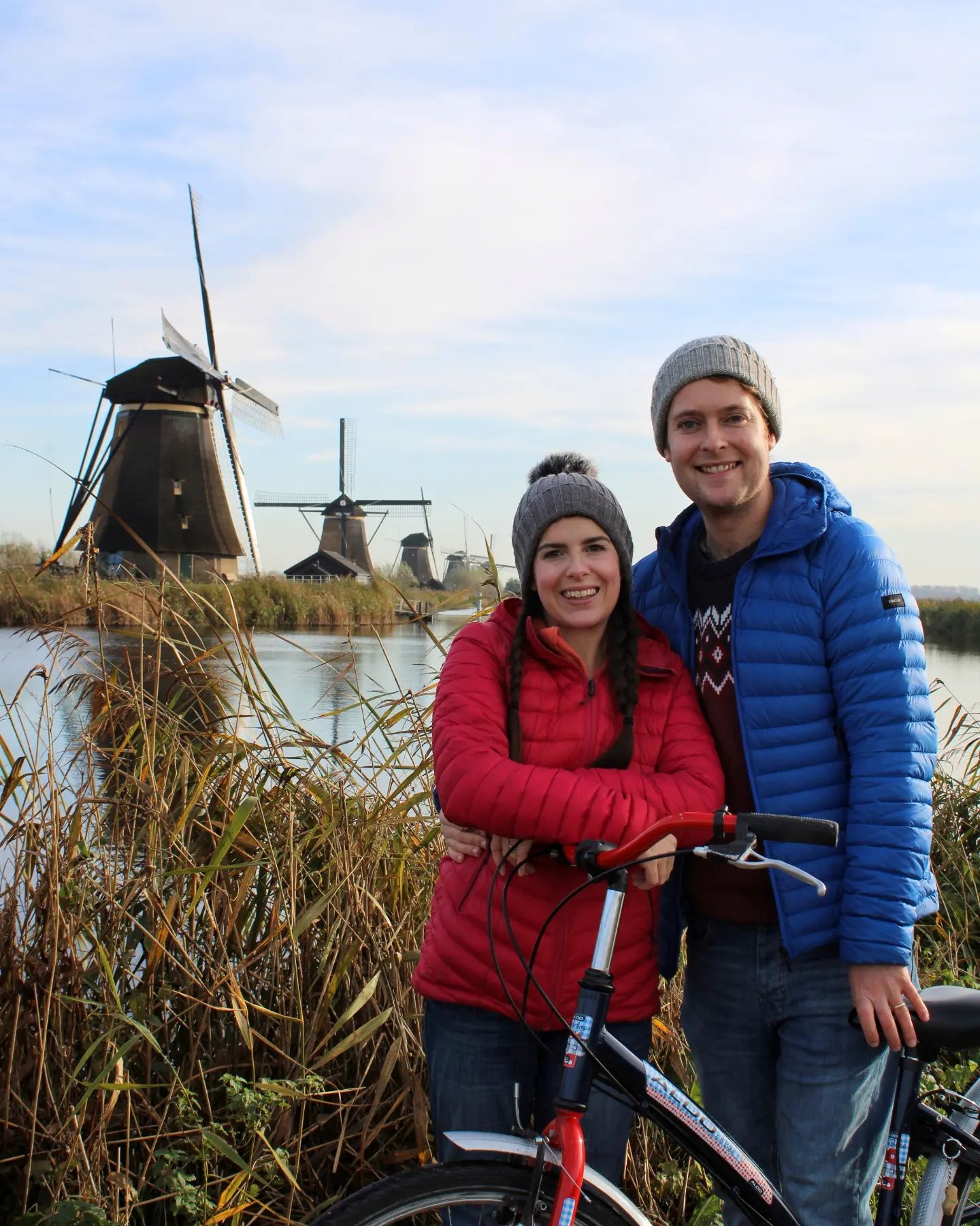a couple wearing puffer coats and wooly hats standing in front of 5 Kinderdijk windmills. The woman is leaning on a bicycle.