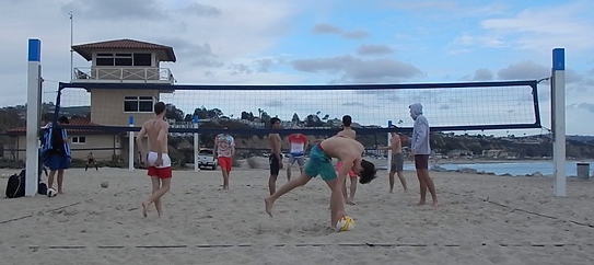 A bunch of guys, some shirtless, one in a hoodie, are playing a game of volleyball on the beach not allowing the cloudy skies to get in their way.
