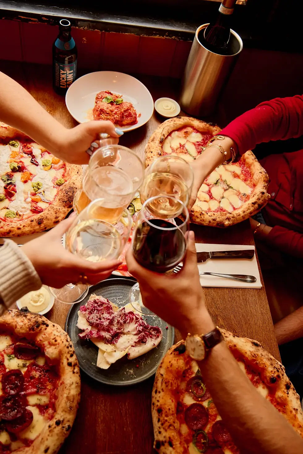 Friends clinking wine glasses while sharing pizza and small plates.