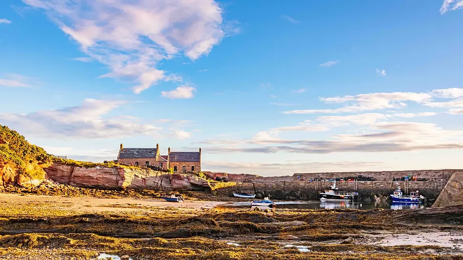 Scottish harbour at low tide with fishing boats, a stone harbour wall and coastal cottages under a blue sky.