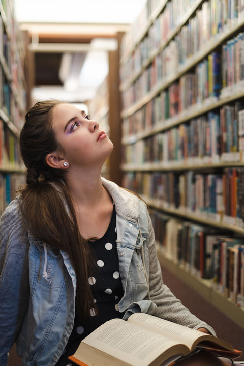 Young woman in library looking up with book, surrounded by bookshelves.