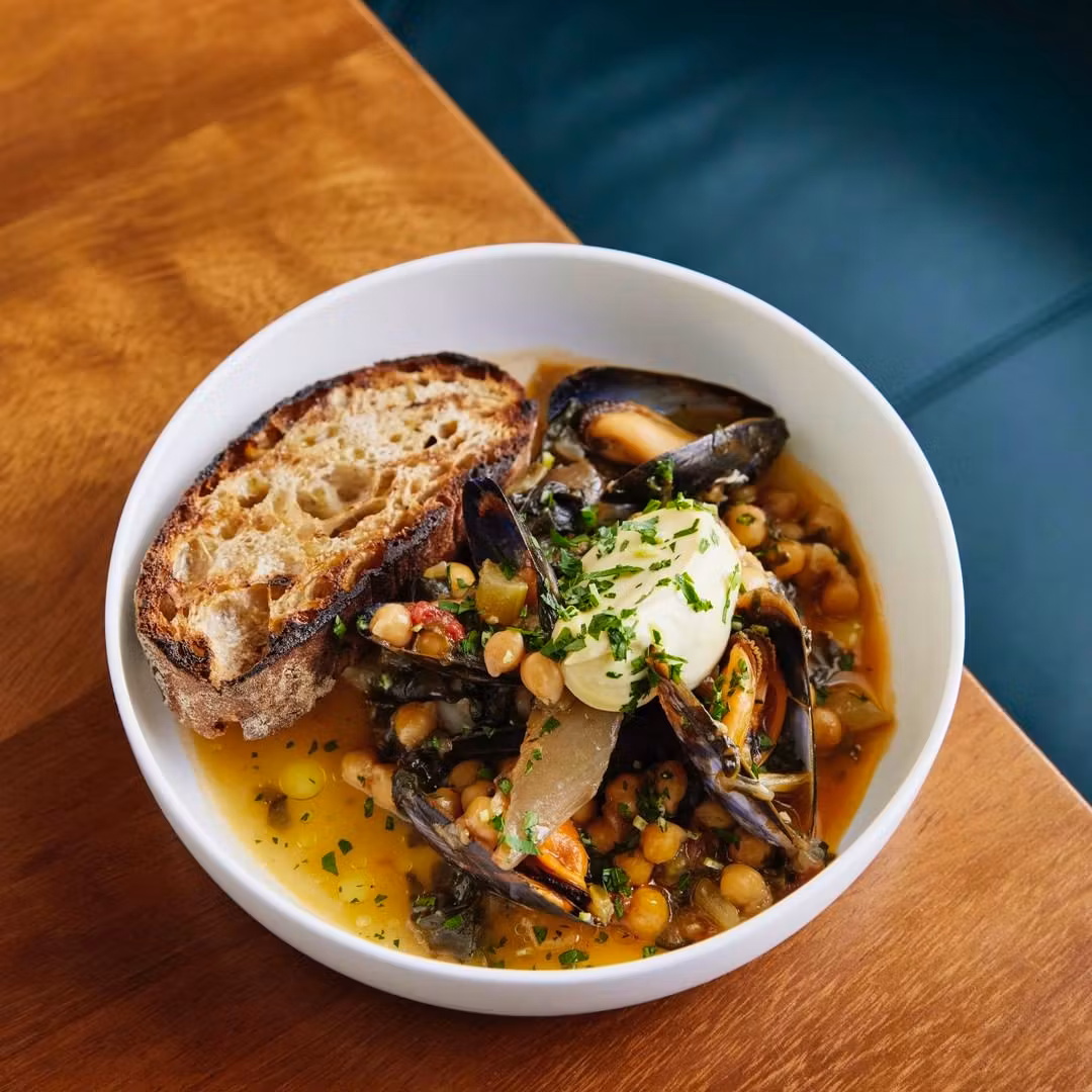 White bowl on a wooden table containing a hearty dish of mussels, chickpeas, and greens in broth, topped with a dollop of cream and parsley, served with a slice of crusty bread.