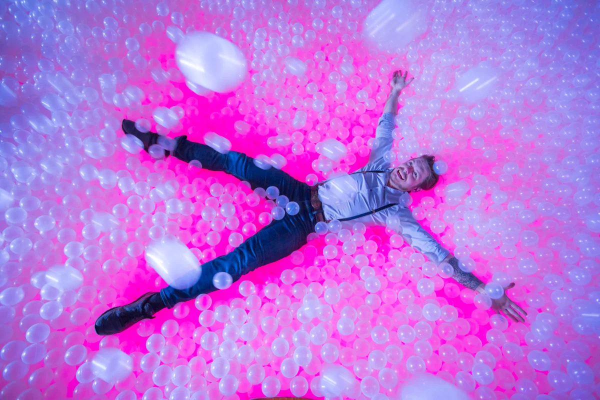 Person lying in a glowing pink ball pit at Ballie Ballerson, arms spread and smiling.