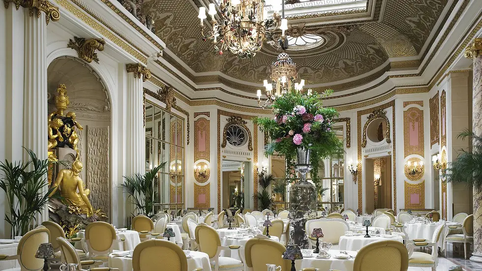A luxurious dining room with ornate chandeliers, floral arrangements, and tables set for afternoon tea, featuring a pianist, at The Palm Court in The Ritz.