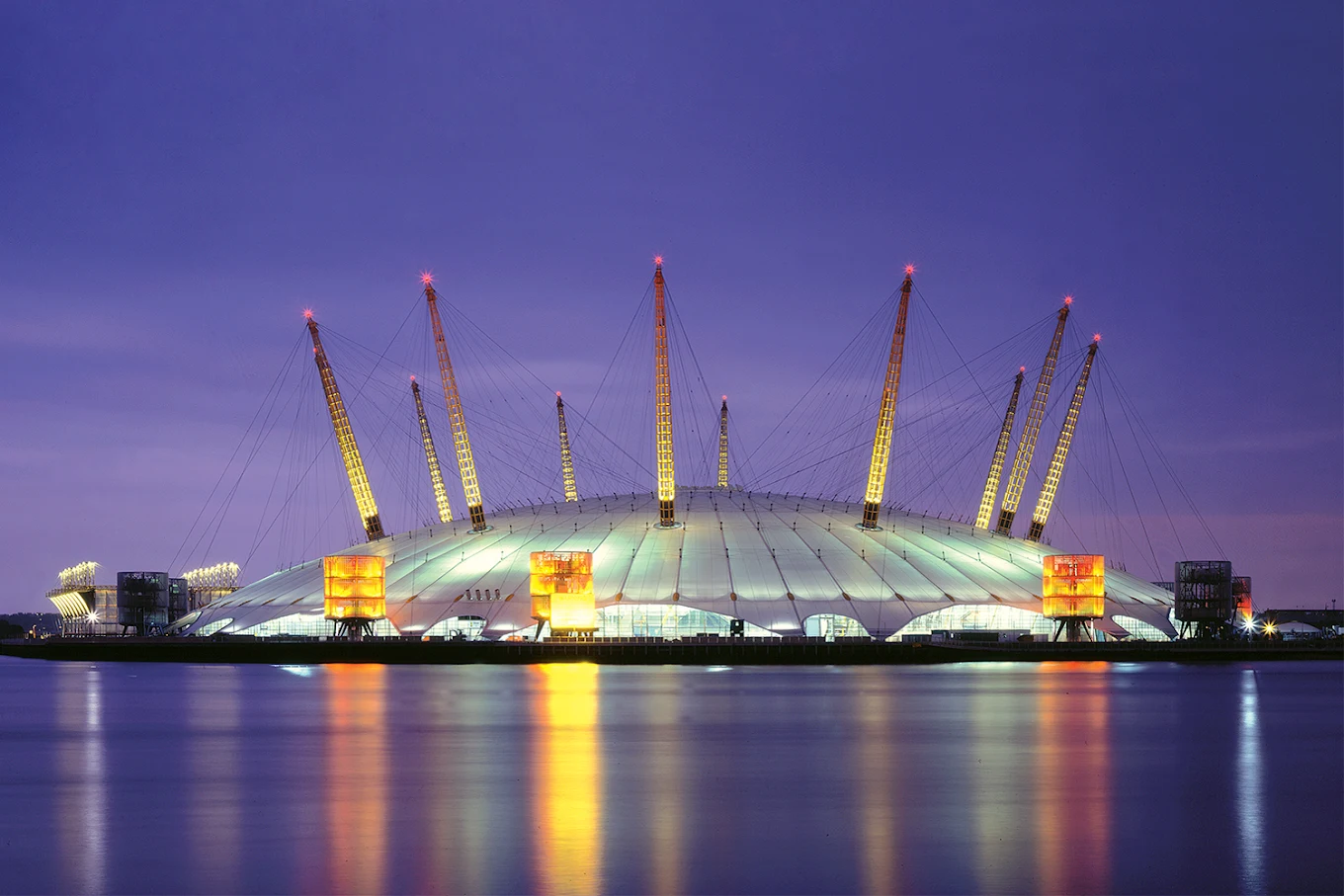 The O2 Arena at night, lit in purple and gold, reflected across the River Thames.