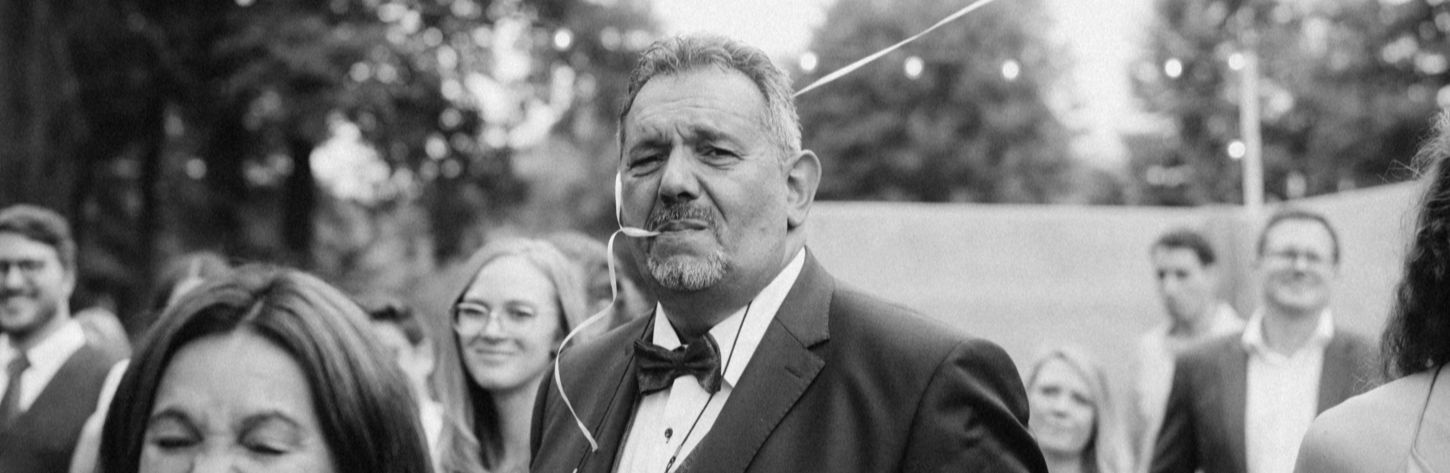 Schnappschuss, Momentaufnahme einer Hochzeitsgesellschaft. Der Brautvater schaut ernst zur Kamera und hat ein Band im Mund an dem ein Luftballong festgemacht ist. Um ihn herum sind viele lachende Menschen. 