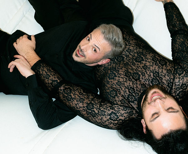 Queer Hochzeitspaar was Schick in Schwarz angezogen ist und auf dem Boden einer Hüpfburg liegt. Der Bräutigam mit hellen Haaren liegt angelehnt auf dem Bauch des Bräutigam mit dunklen, langen Haaren welcher lacht. Sie spielen mit ihren Händen.