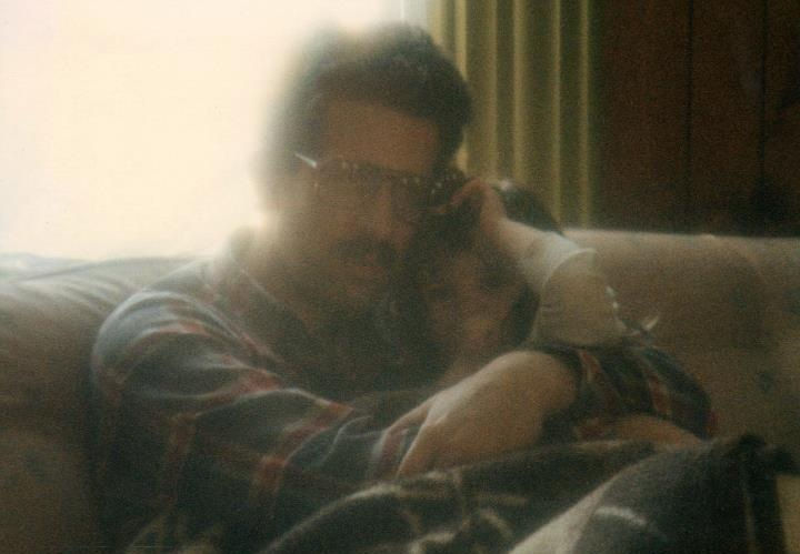 Man in glasses and a plaid shirt hugs a child on a couch. The room is softly lit, creating a cozy and warm atmosphere.
