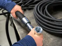 Close up of a hydraulic hose assemble in a workshop, with hands aligning a metal threaded fitting onto a heavy duty hydraulic hose beside coiled hydraulic hose lines on a steel workbench.