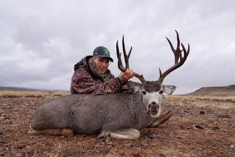 A hunter sits next to his trophy mule deer with grey skies behind him.